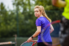 Frau in violettem Shirt mit Tennisracket auf einem Tennisplatz, im Hintergrund Bäume.