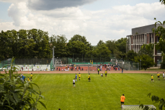 Fußballplatz mit Spielern, Zuschauern am Rand, Toren, Bäumen und bewölktem Himmel.