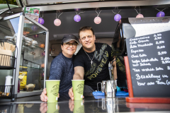 Zwei Personen hinter einer Kaffeetheke, davor zwei grüne Becher und rechts eine Getränketafel.