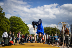 Mann in blauem Shirt macht einen Handstand auf einem gelben Balken, umringt von Zuschauern im Freien.