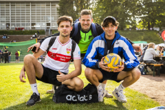 Drei Männer posieren auf einer Wiese; einer hält einen Fußball, vor ihnen eine schwarze Adidas-Tasche.