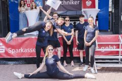 Gruppe von Sportlern in blauen Shirts vor einer Bühne, zwei Frauen machen einen Spagat.