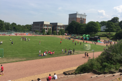 Sportplatz mit vielen Personen, Gebäuden, Bäumen und einem Fußballtor unter blauem Himmel.