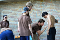 Mehrere Personen vor einer Backsteinwand; eine bemalt den Oberkörper eines Mannes mit pinkem Stift.