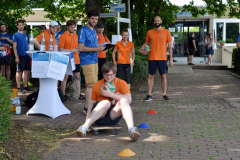 Personen mit Sportshirts stehen und sitzen vor einem Gebäude; einige halten Bälle, davor Hütchen auf dem Boden.