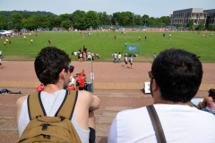 Zwei Männer mit Rucksack schauen auf ein Fußballfeld mit vielen Spielern und Zuschauern.