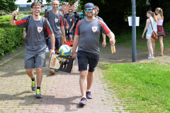 Mehrere Männer in grauen Sportshirts gehen auf einem Weg; zwei tragen Getränkekisten und Flaschen.