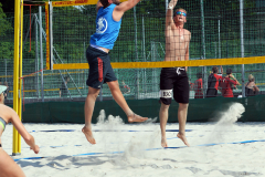 Zwei Männer springen am Netz im Sand, ein Volleyball fliegt darüber.