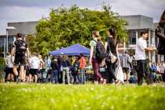 Menschenversammlung auf einem Campus mit blauem Zelt und Bäumen im Hintergrund.