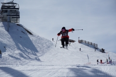 Skifahrer springt auf einer verschneiten Piste vor einer Bergseilbahn.