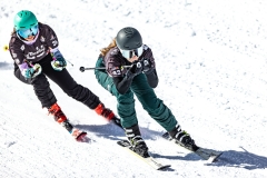 Zwei Skifahrerinnen in Rennausrüstung fahren auf einer verschneiten Piste nebeneinander.