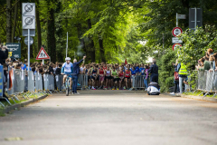 Startlinie eines Straßenlaufs mit vielen Läufern, Absperrgittern, Zuschauern, Fahrradfahrer und Verkehrsschildern im Grünen.